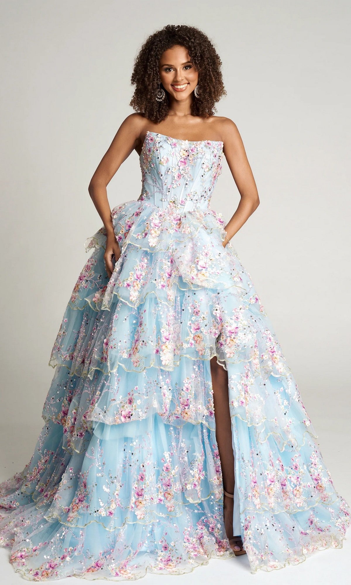 A woman smiles in the Portia and Scarlett PS26051 strapless floral ball gown in blue, featuring tiered layers, floral appliqué details, and a high slit, set against a plain background.